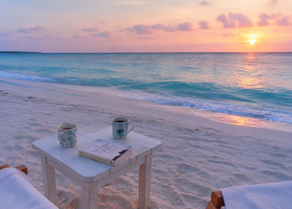 Coffee cups and a paperback on white sand at sunrise